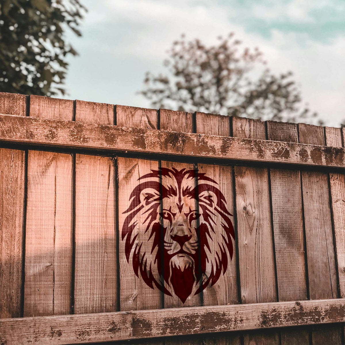 A rustic wood fence displays the Regal Lion Head Stencil in a rich dark red finish.