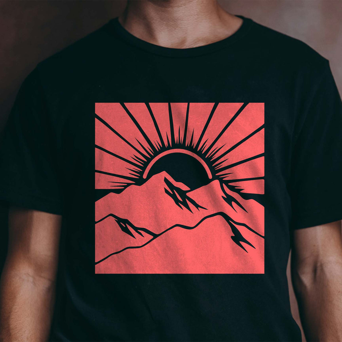 A coral-red Sunrise Mountain Landscape Stencil displays rugged peaks and dramatic sunrays on a black t-shirt.