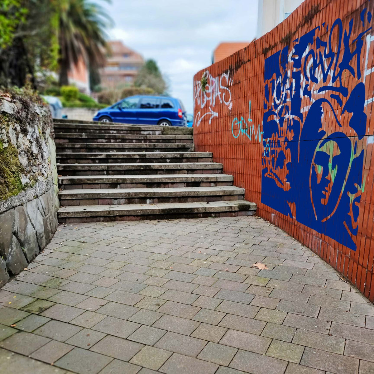 Bold blue Urban Street Graffiti Stencil artwork brings layered tags and a hooded face to a brick wall.