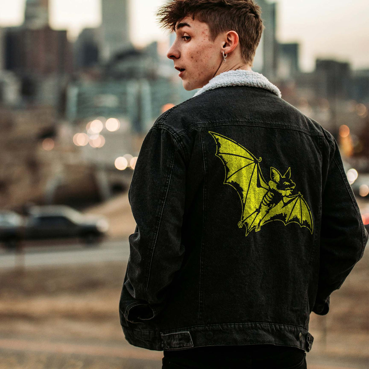 Flying Bat Stencil featured in yellow on the back of a black jacket in a city scene.