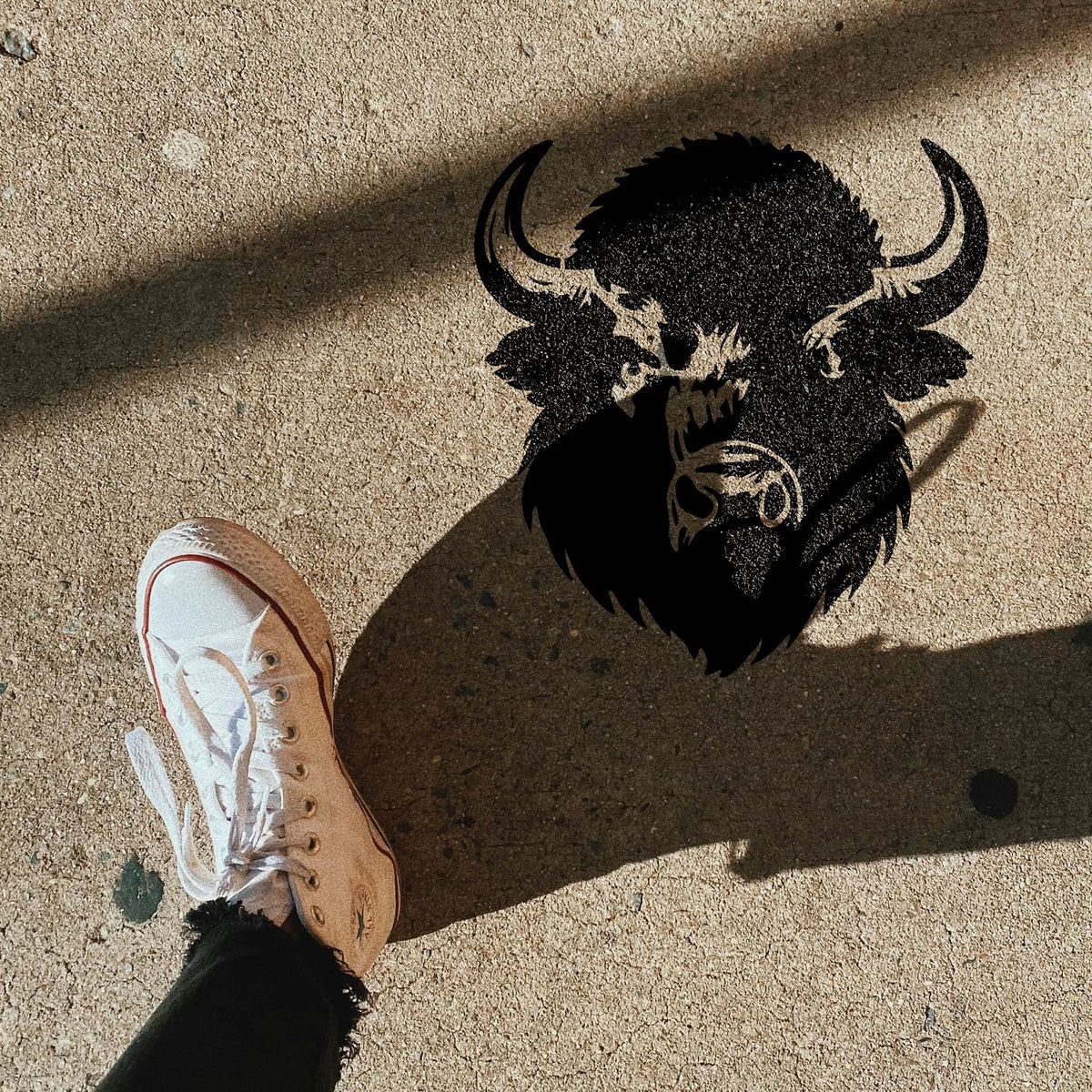 Shaggy Buffalo Head Stencil painted black on concrete pavement beside a white sneaker and strong shadow.