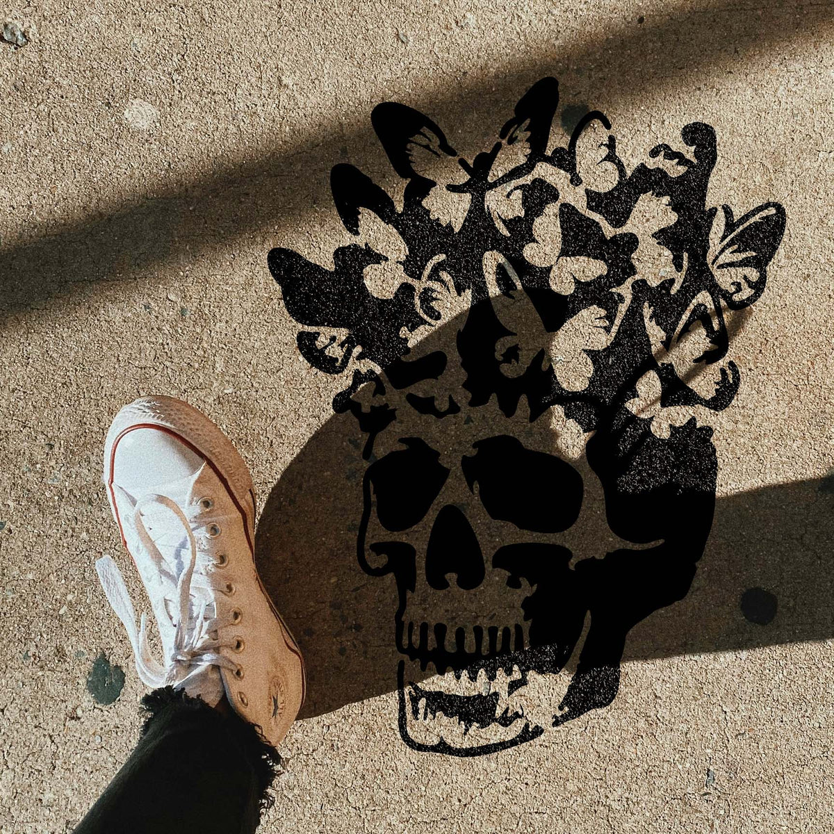 Butterfly Bloom Skull Stencil stands out on pavement beside a sneaker in dramatic sunlight