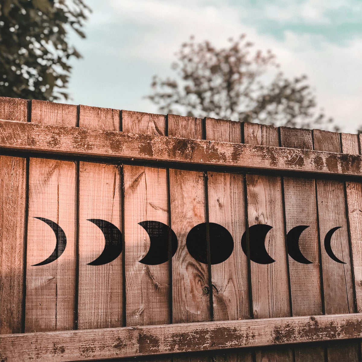 Seven moon phases from the Celestial Moon Phase Border Stencil span a rustic wooden fence in black.