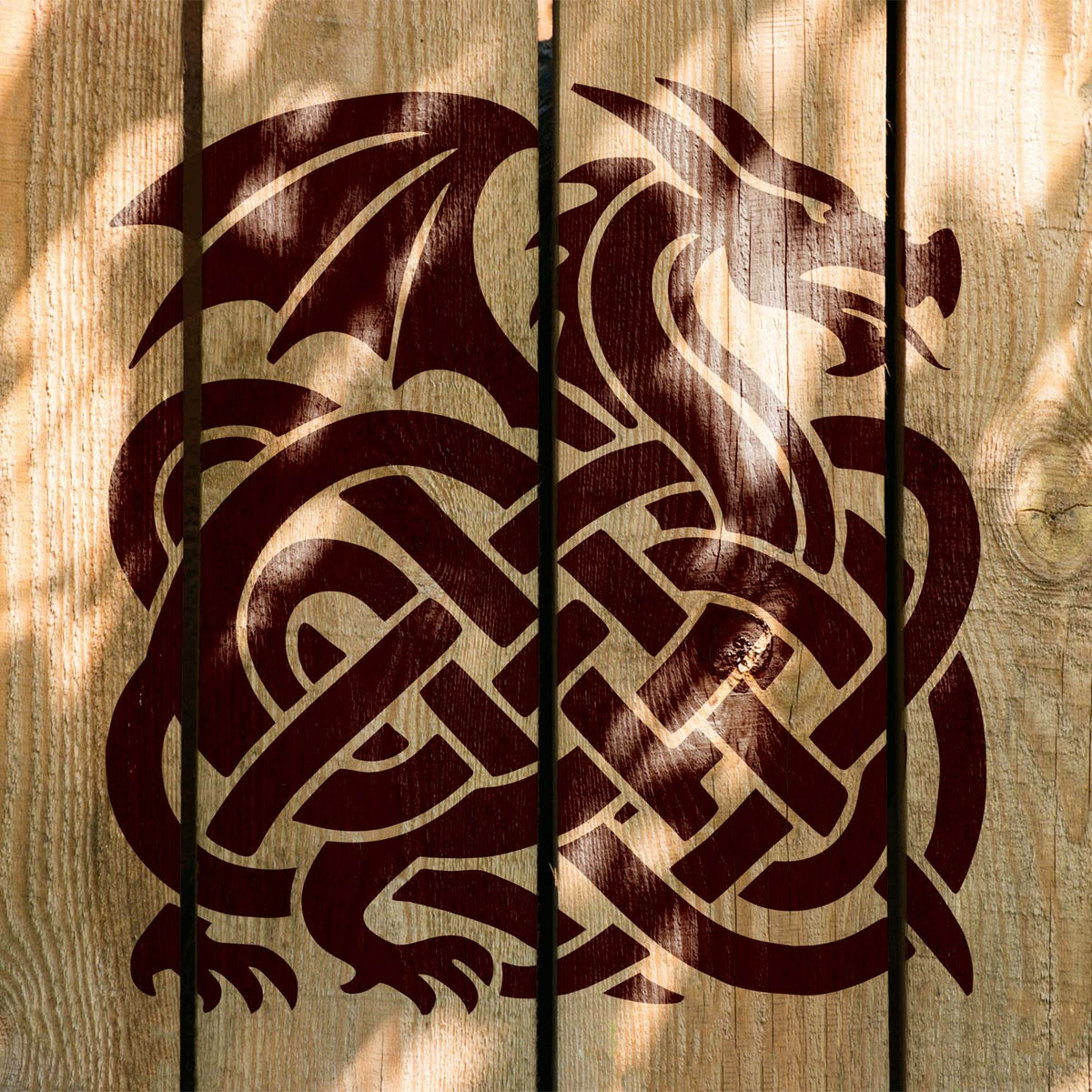 Sunlit wooden planks showcase the Celtic Dragon Knot Stencil with sweeping wings and an interlaced dragon body.