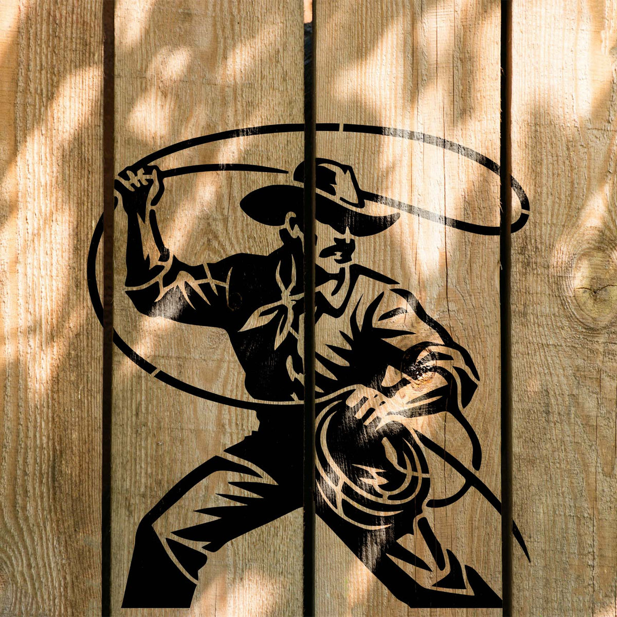 Cowboy Lasso Action Stencil painted in black across sunlit vertical wooden fence boards.