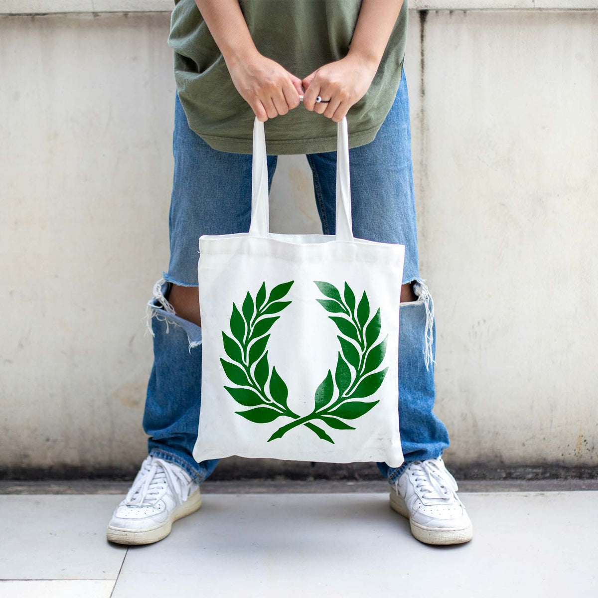 A white tote bag features the Elegant Laurel Wreath Stencil in green, held against ripped blue jeans.