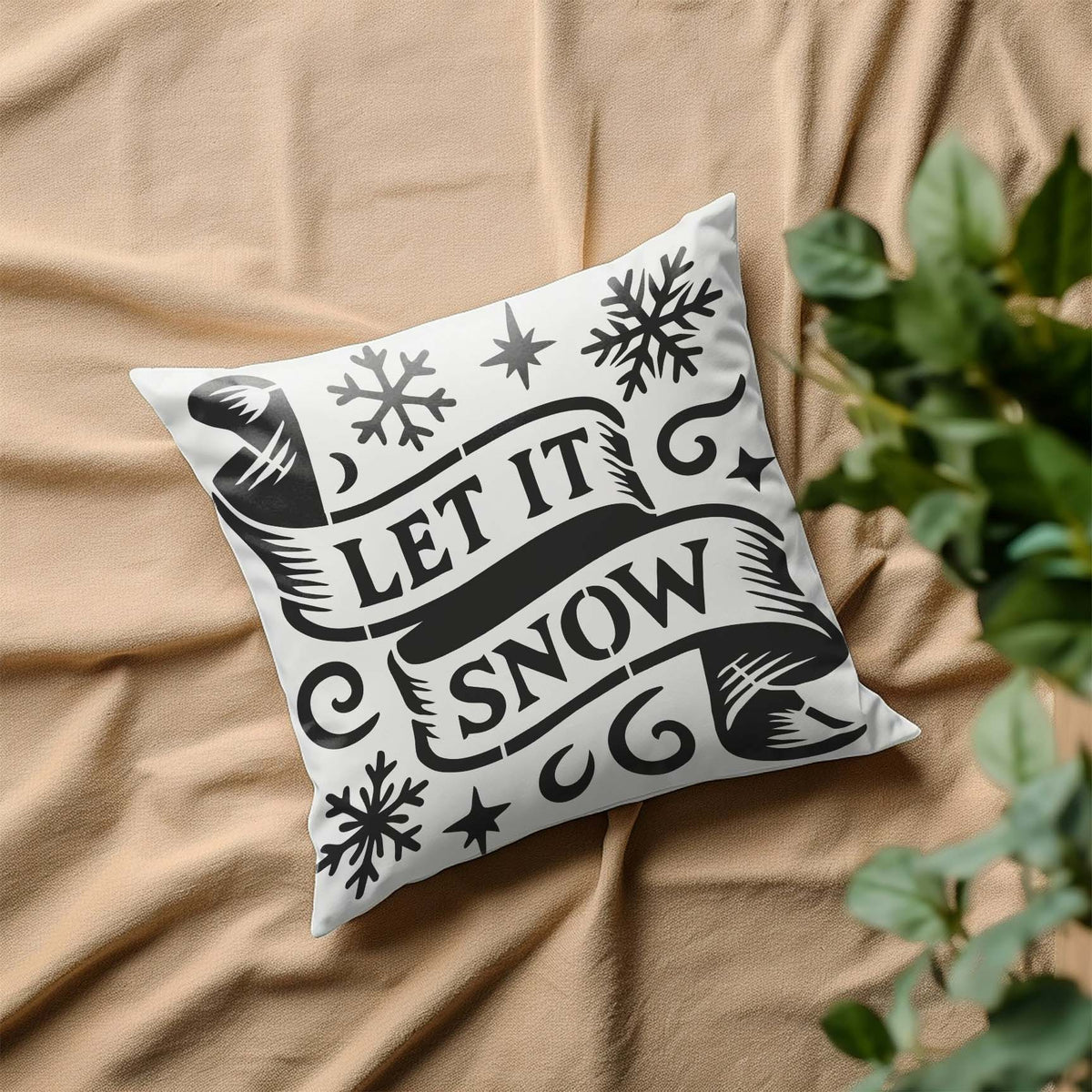 A white pillow displays the Let It Snow Banner Stencil with sweeping ribbons and winter snowflakes.