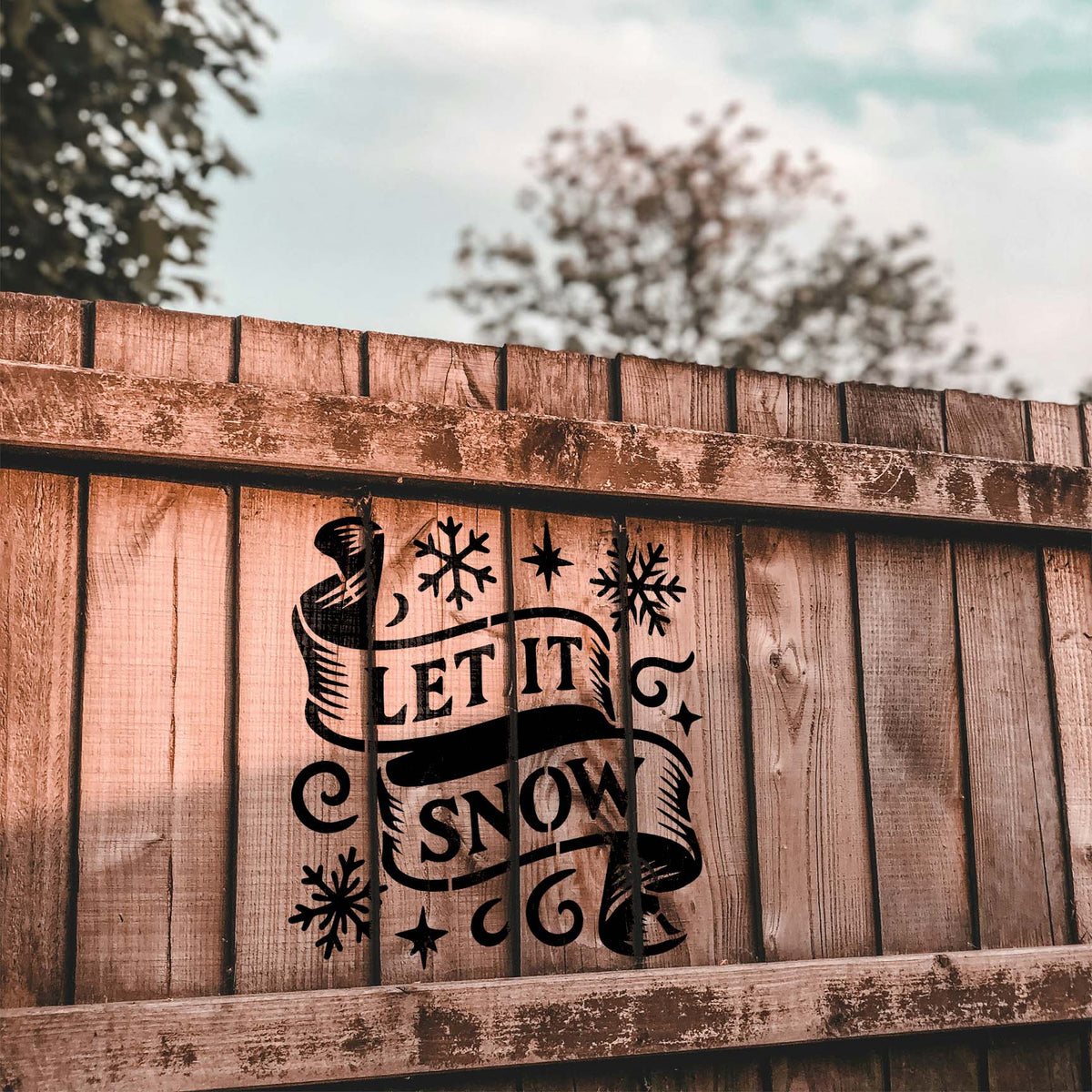 Let It Snow Banner Stencil brings bold ribbon lettering and snowflakes to a rustic wooden fence.