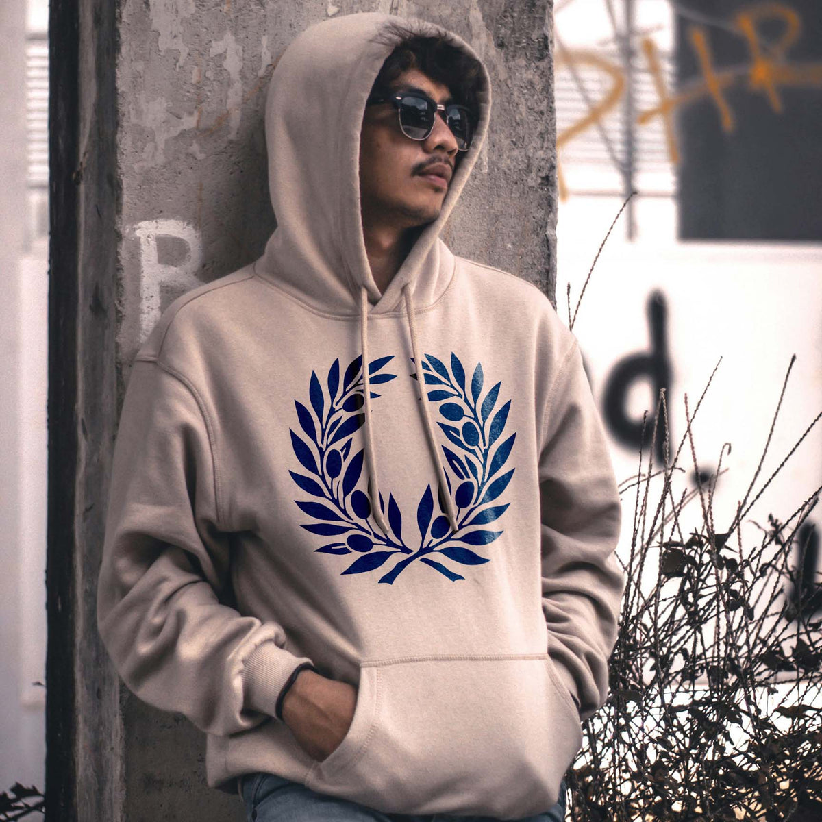 A bold blue Olive Branch Wreath Stencil adorns the front of a beige hoodie with leafy branches and olives.