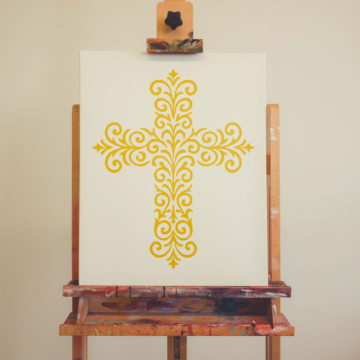 Displayed on an easel, the Ornate Filigree Cross Stencil forms an elegant gold design on white canvas