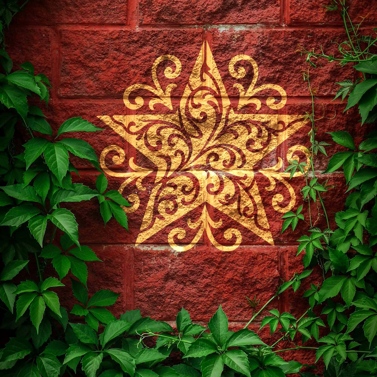 Framed by green vines, the Ornate Filigree Star Stencil gleams gold on a red brick wall