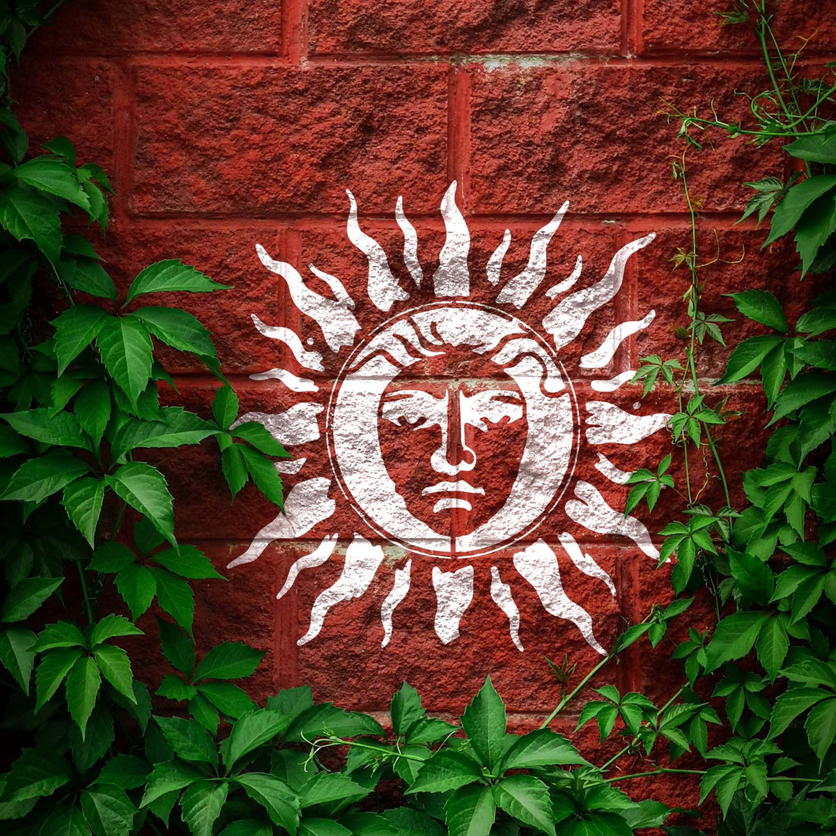 Radiant Sun Face Stencil shines in white across a red brick wall framed by green vines