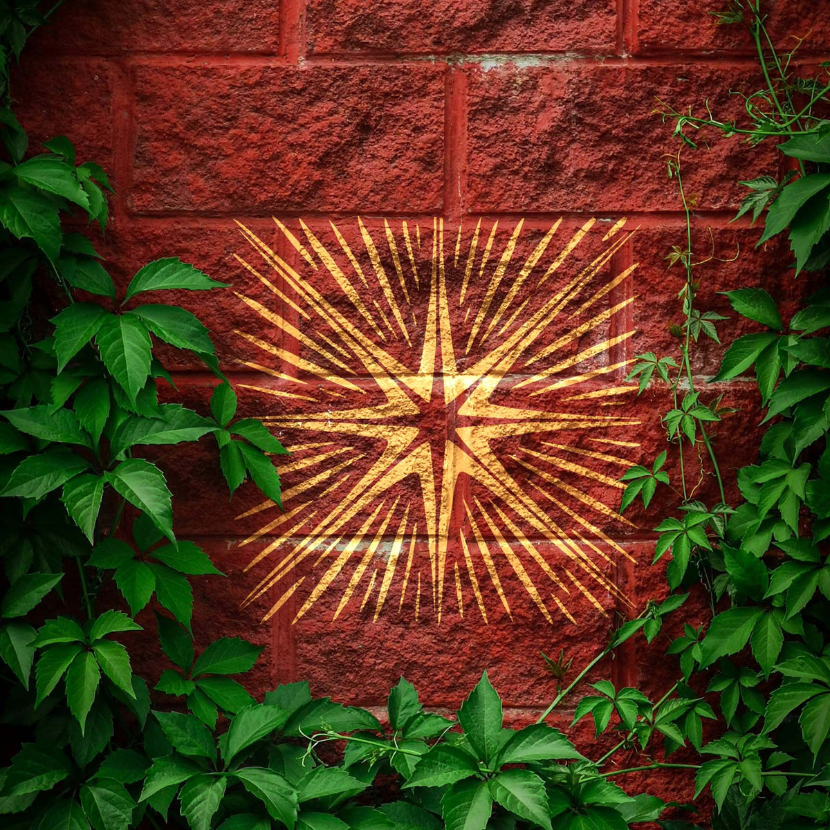 Golden Radiating Eight-Point Star Burst Stencil glows across a red brick wall framed by leafy vines
