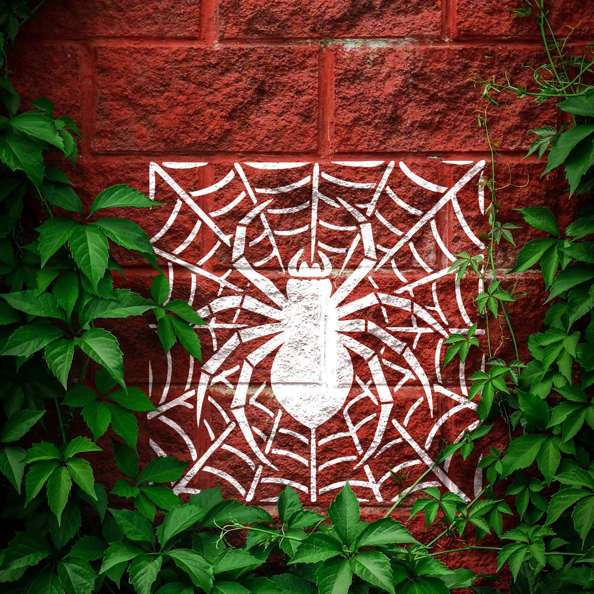 Spider Web Silhouette Stencil reveals a bold white spider and web across a leafy red brick wall