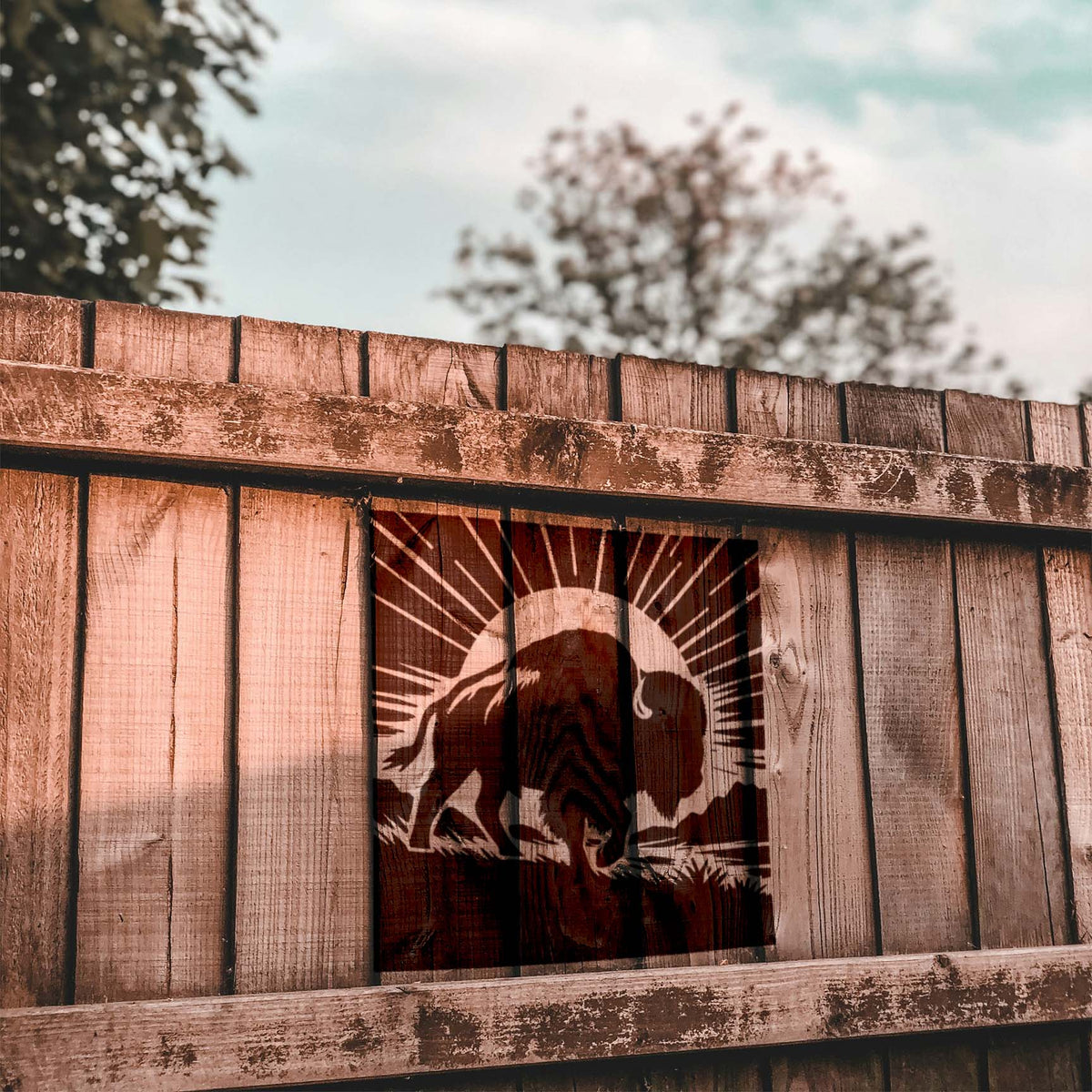 Rustic wooden fence displays the Sunrise Buffalo Silhouette Stencil with a bison, grassy plains, and sun rays.