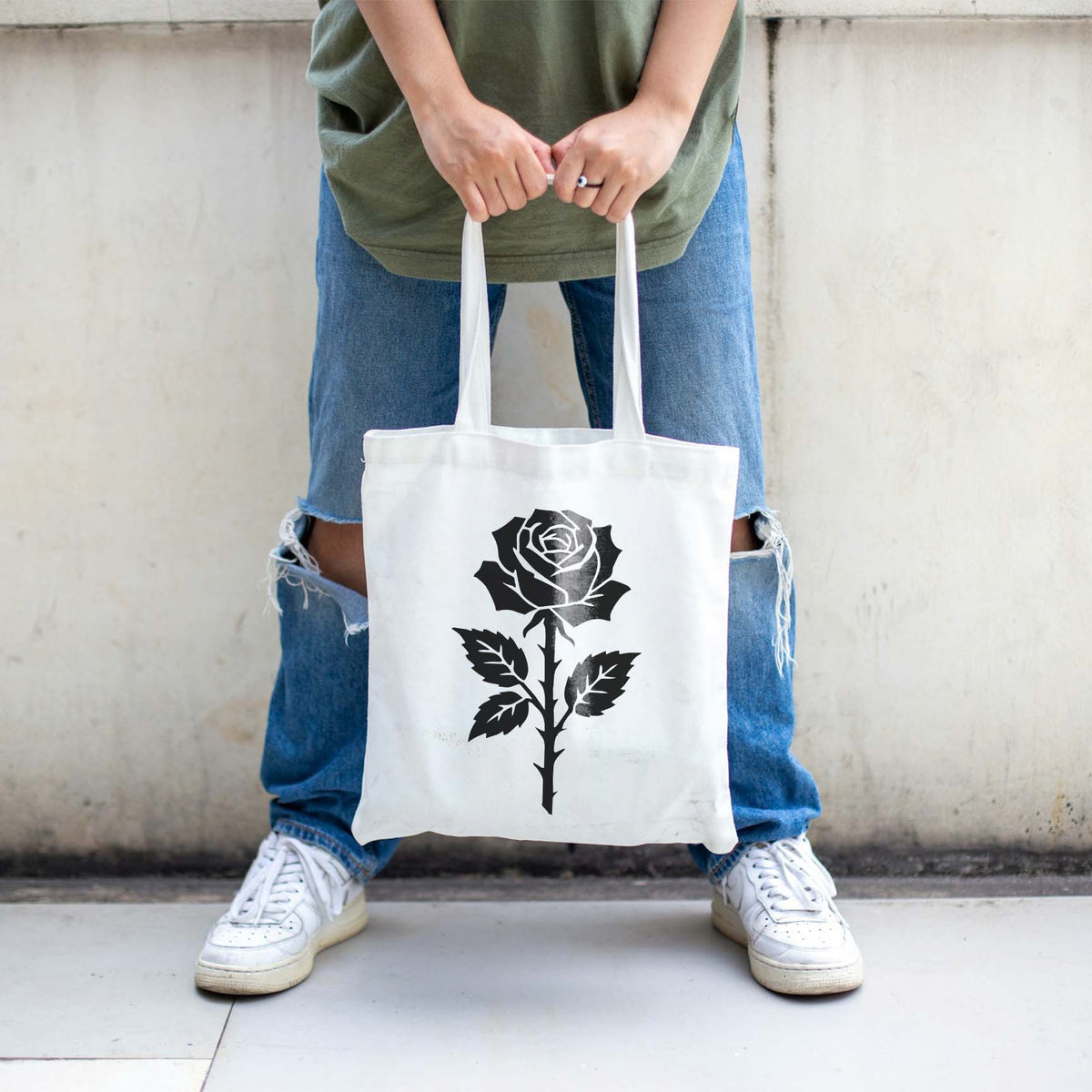 White tote bag featuring the Thorned Rose Blossom Stencil in black, held in front of ripped jeans.
