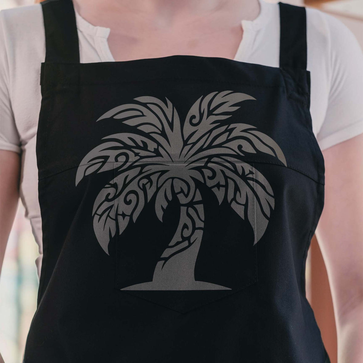 Silver Tribal Palm Tree Stencil creates swirling tropical fronds across a black apron