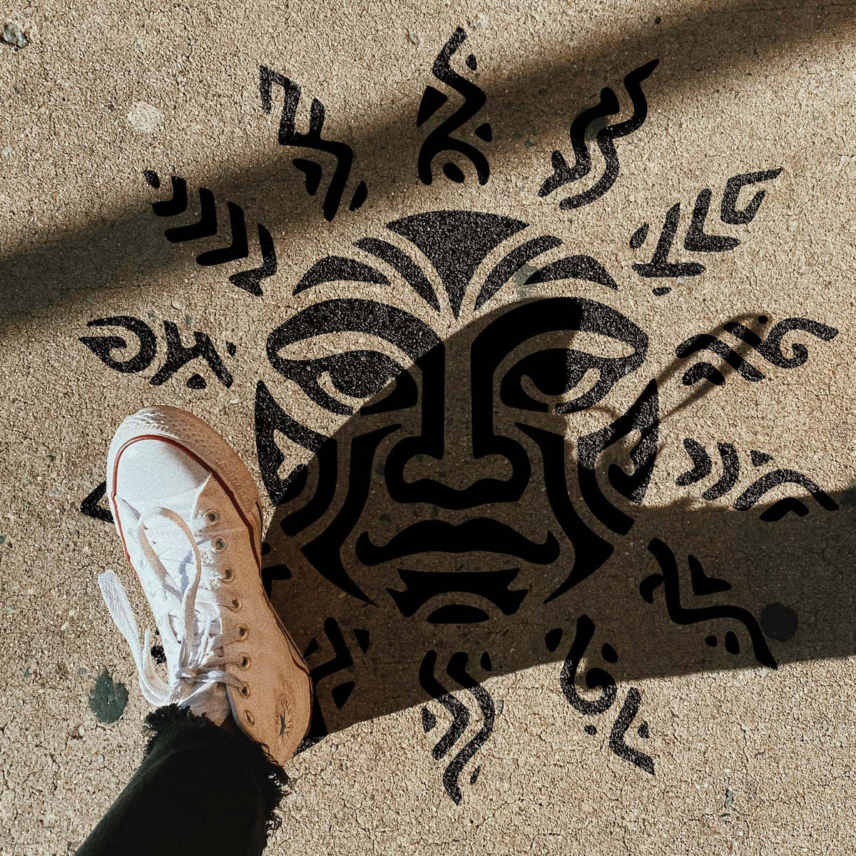 Street-style Tribal Sun Face Stencil appears beneath dramatic shadows on a concrete sidewalk