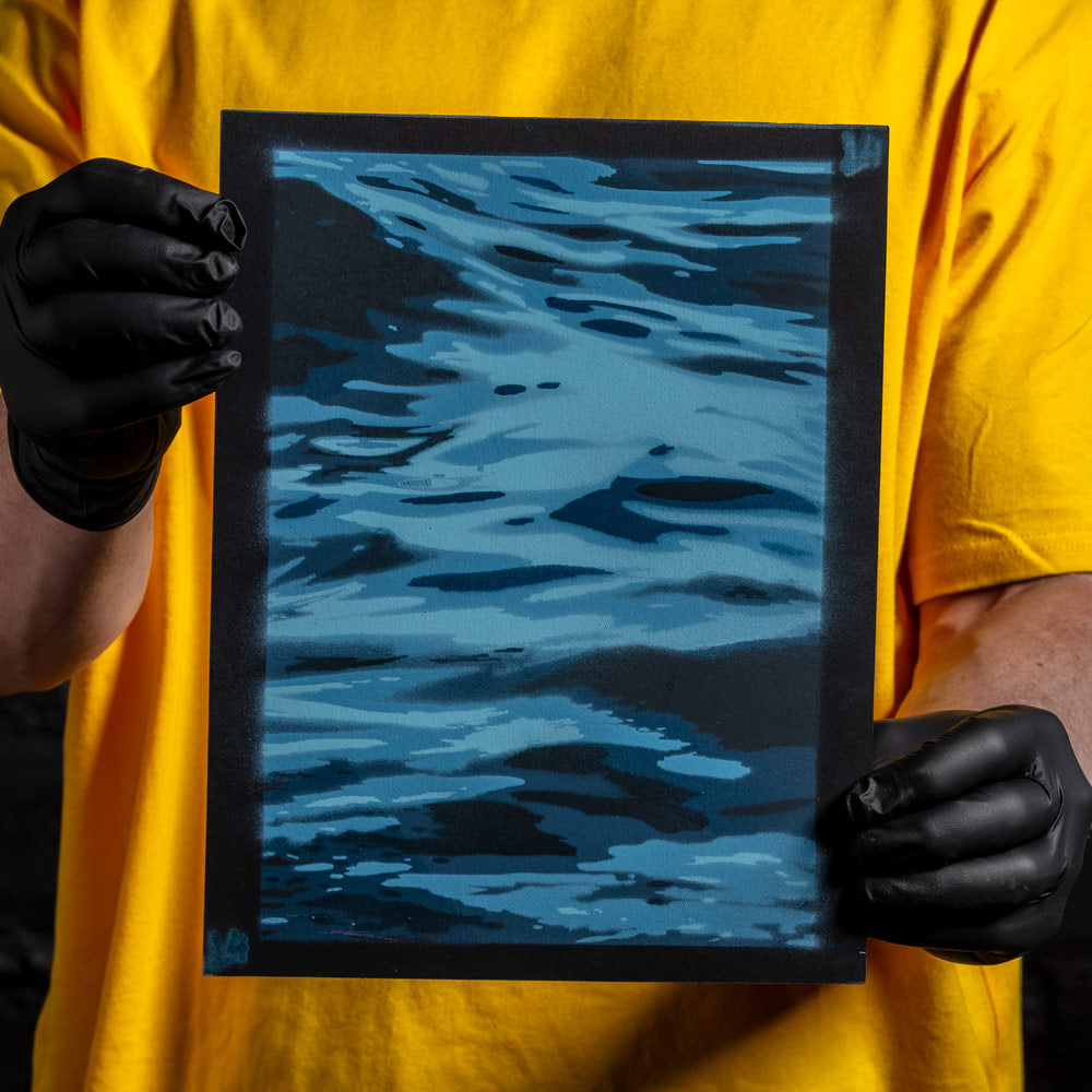 Water reflection layered stencil artwork in blue tones spray paint held by artist wearing black gloves