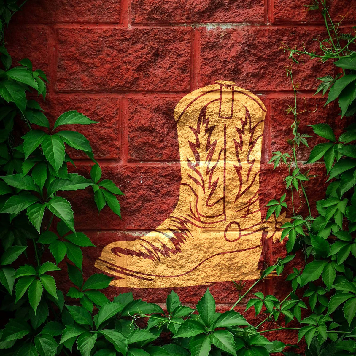 Western Cowboy Boot with Spur Stencil is shown in gold on a red brick wall framed by ivy.