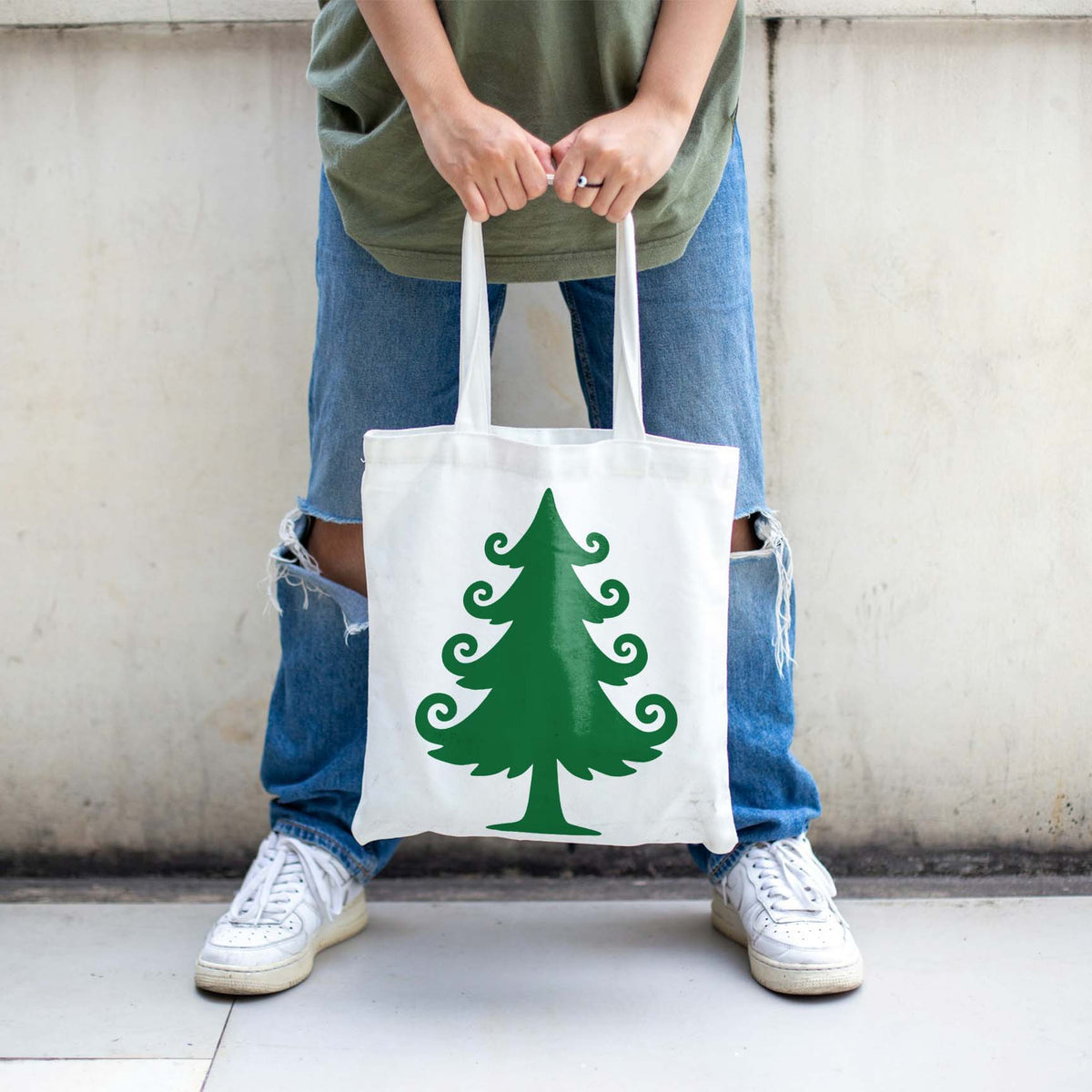 White tote bag featuring the Whimsical Christmas Tree Stencil in green, held between ripped jeans.