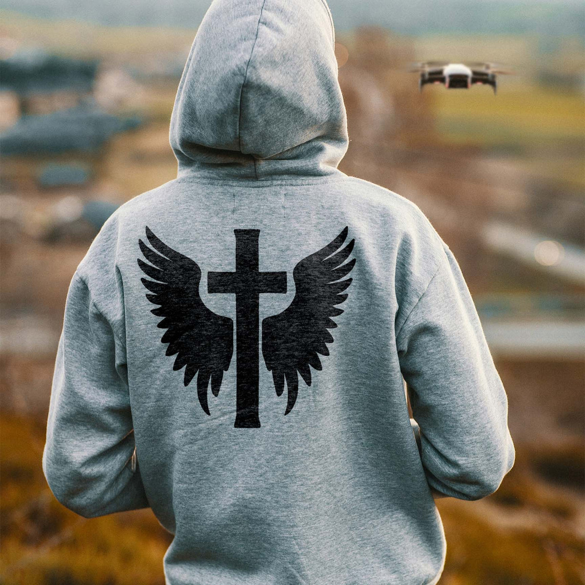 A gray hoodie features Winged Christian Cross Stencil printed boldly across the back outdoors