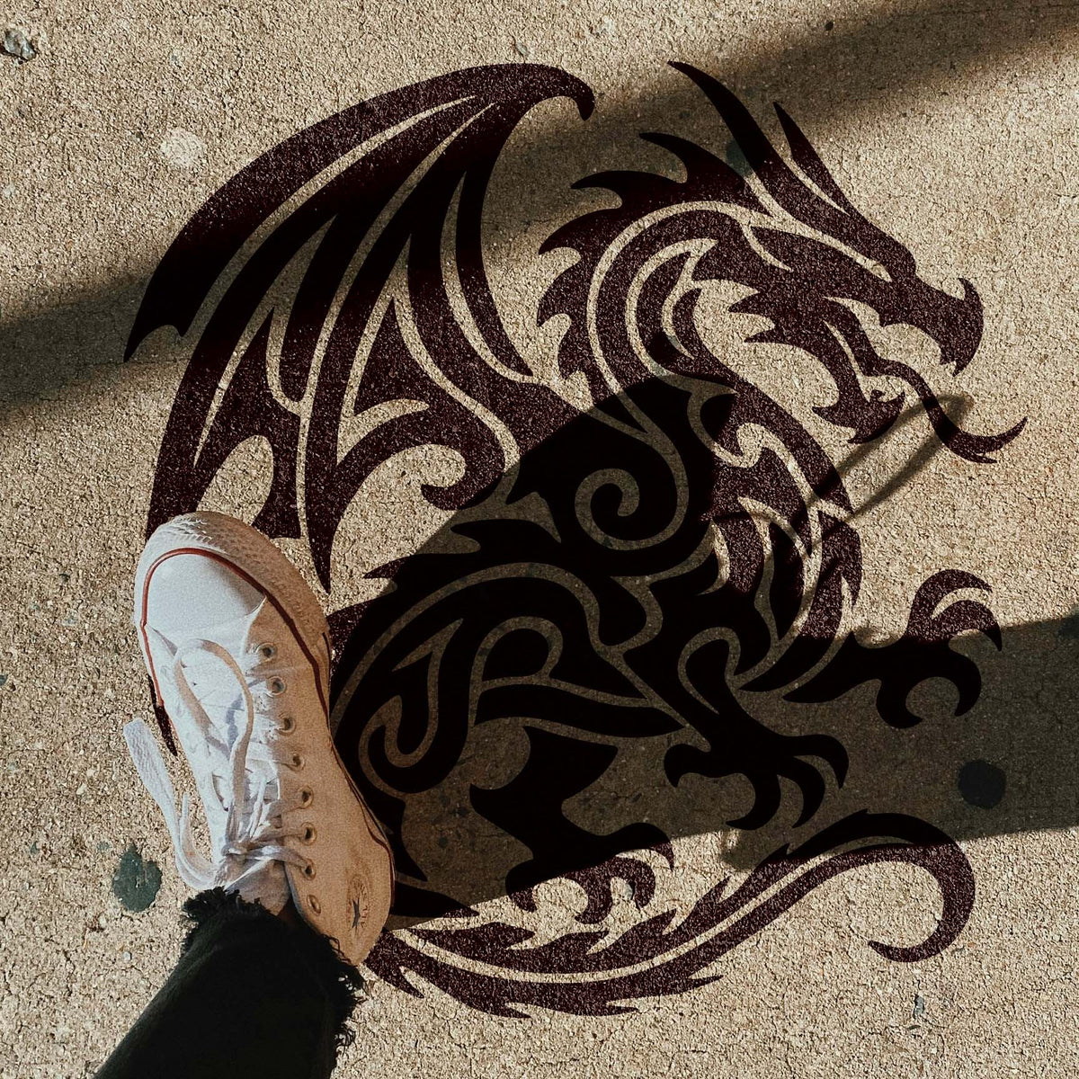 Intricate swirls shape the Tribal Fire Dragon Stencil across a sunlit concrete sidewalk beside a white sneaker.