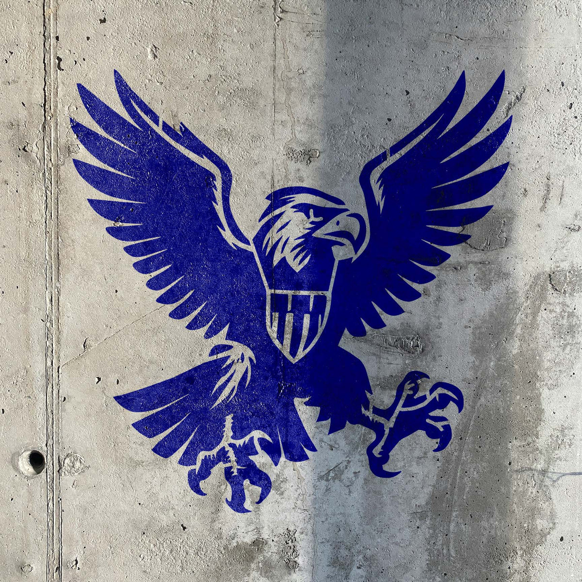 A rugged concrete wall showcases the American Eagle Crest Stencil in vivid blue with a shielded eagle.
