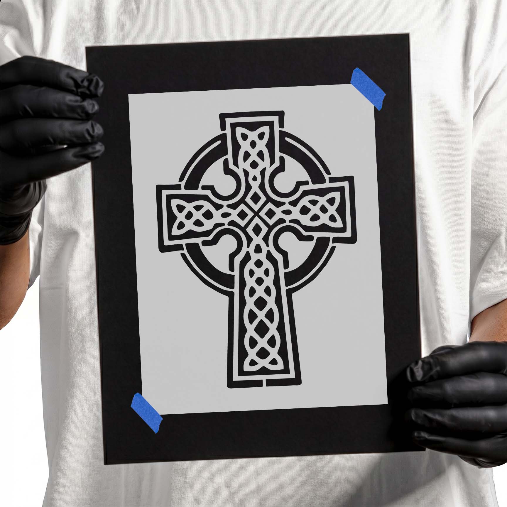 Gloved hands holding Celtic Knot Cross Stencil taped to black cardboard
