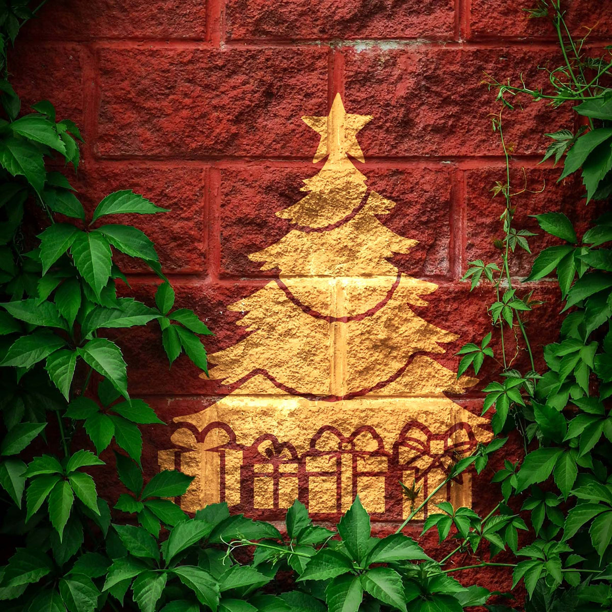 The Christmas Tree with Presents Stencil appears in gold on a red brick wall framed by green vines.