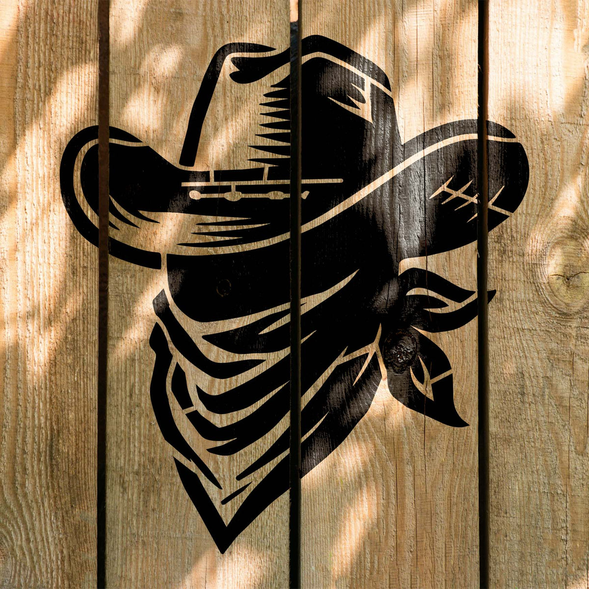 Sunlight highlights the Cowboy Hat & Bandana Stencil painted black across rustic wooden fence planks.