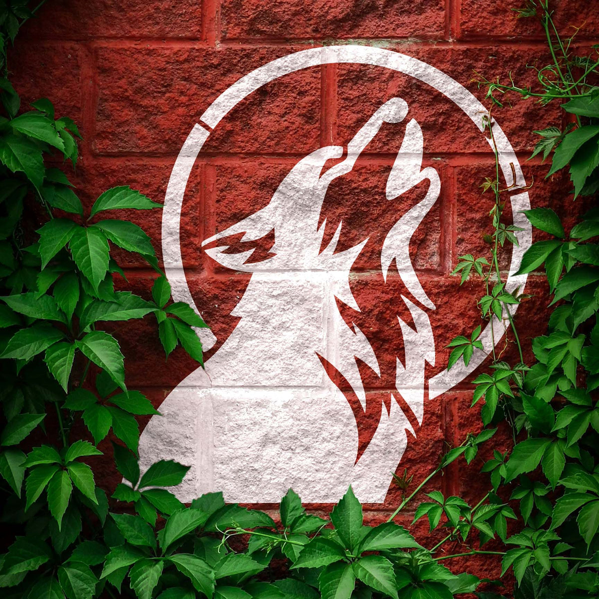 Dog Howling at Moon Stencil painted in white on a red brick wall framed by green ivy.