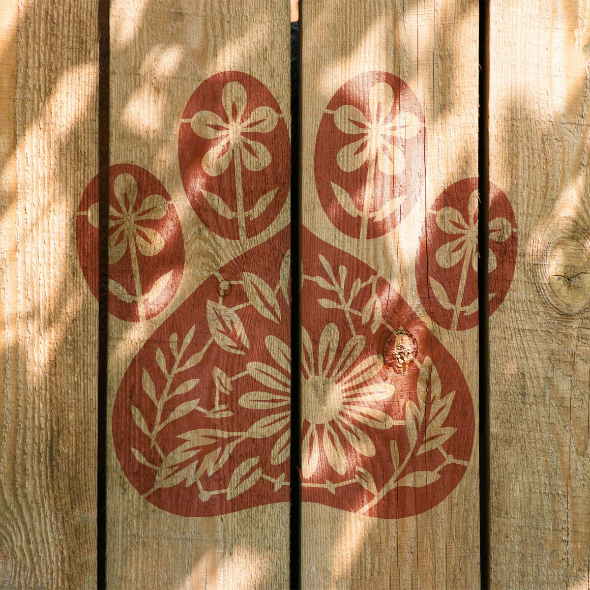 Sunlight crosses the Floral Dog Paw Print Stencil painted in warm red on rustic wood planks