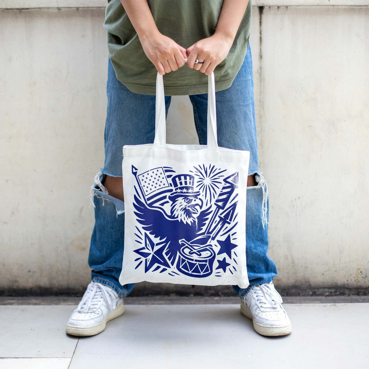 A white tote bag showcases the Fourth of July Eagle Celebration Stencil in blue with a flag and festive drum.