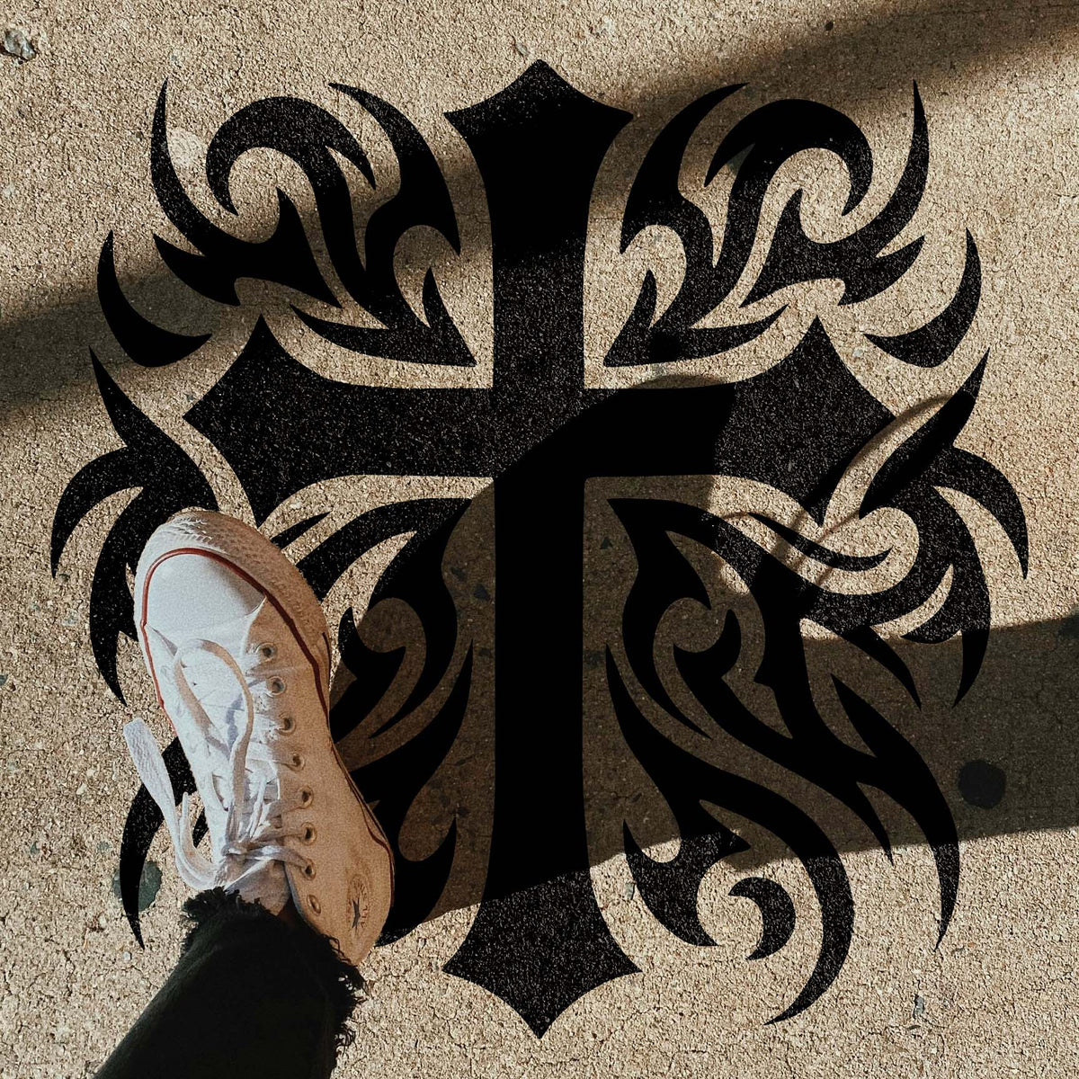Heritage Tribal Cross Stencil shown on pavement beside a white sneaker in sunlight.