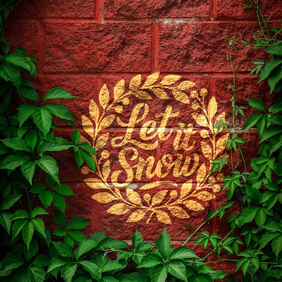 Golden Let It Snow Holiday Wreath Stencil stands out on a red brick wall framed by green vines.