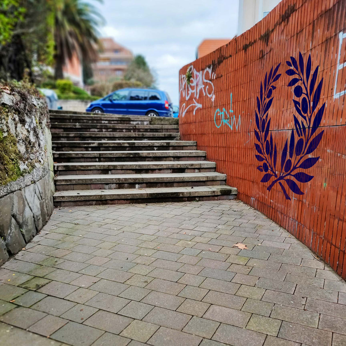 Olive Branch Wreath Stencil painted in deep blue on a red brick wall beside an outdoor staircase.