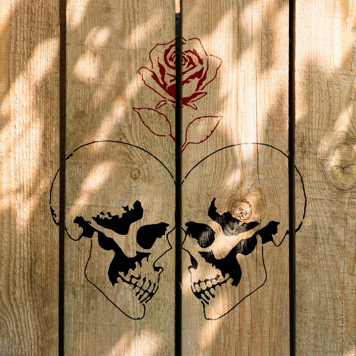 Rustic wood showcases the Rose Between Twin Skulls Stencil framed by sunlight and shadow