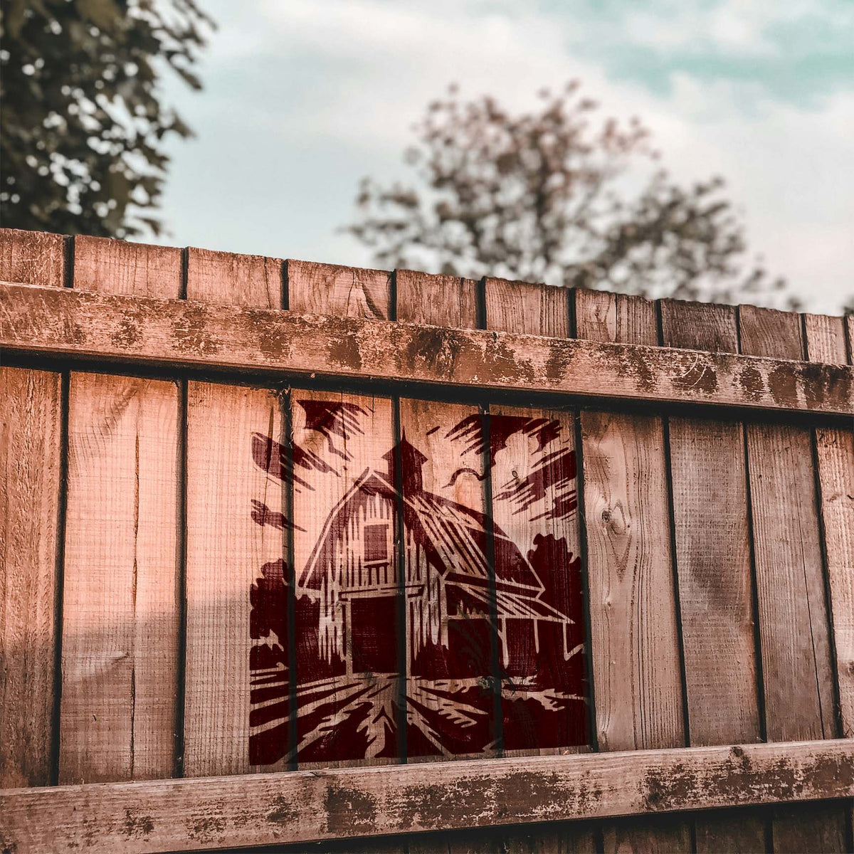 Farmhouse scenery from the Rustic Country Barn Stencil stands out across a rustic wood fence