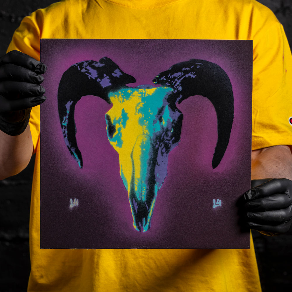 Ram skull layered stencil artwork in purple, teal, and yellow spray paint held by artist wearing black gloves
