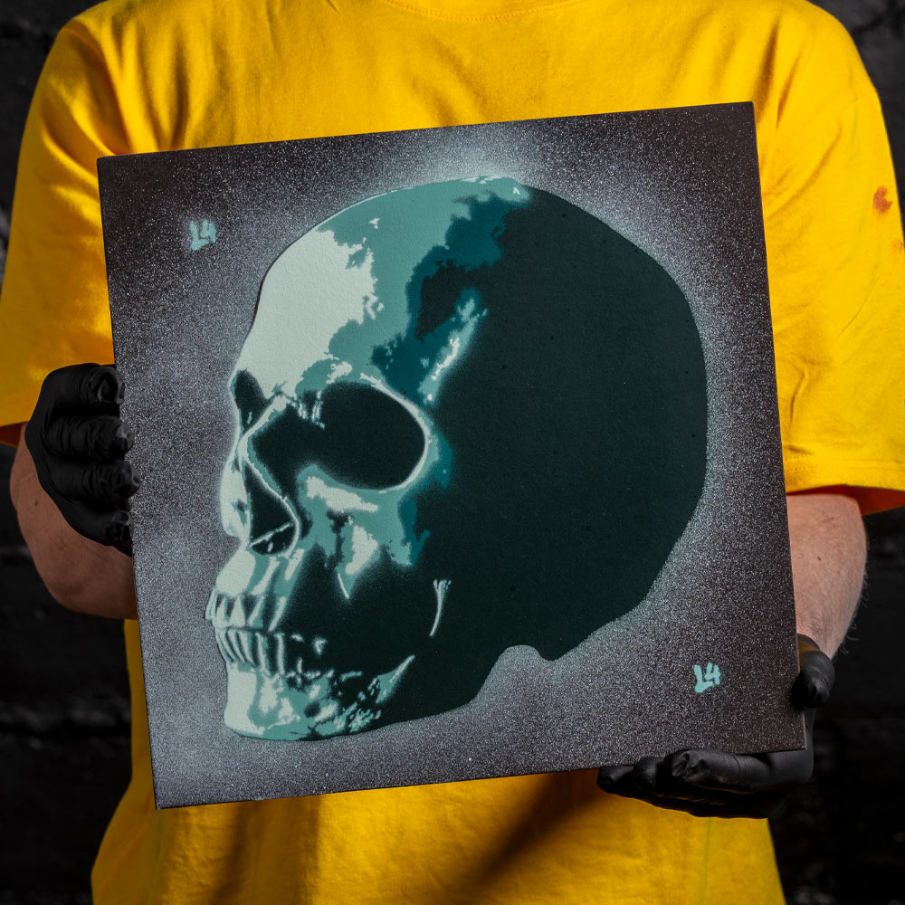 Human skull layered stencil artwork in teal and white spray paint held by artist wearing black gloves