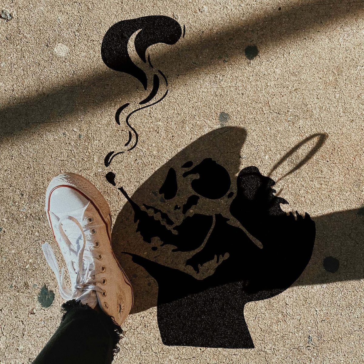 Street-style Smoking Skull Silhouette Stencil appears beside a sneaker on a sunlit sidewalk