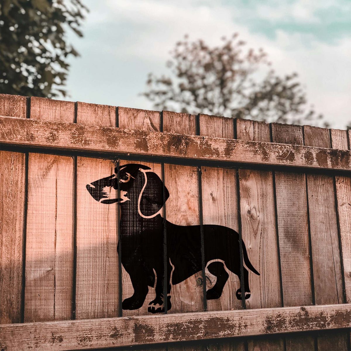 Standing Dachshund Stencil shown in black across weathered wooden fence boards.