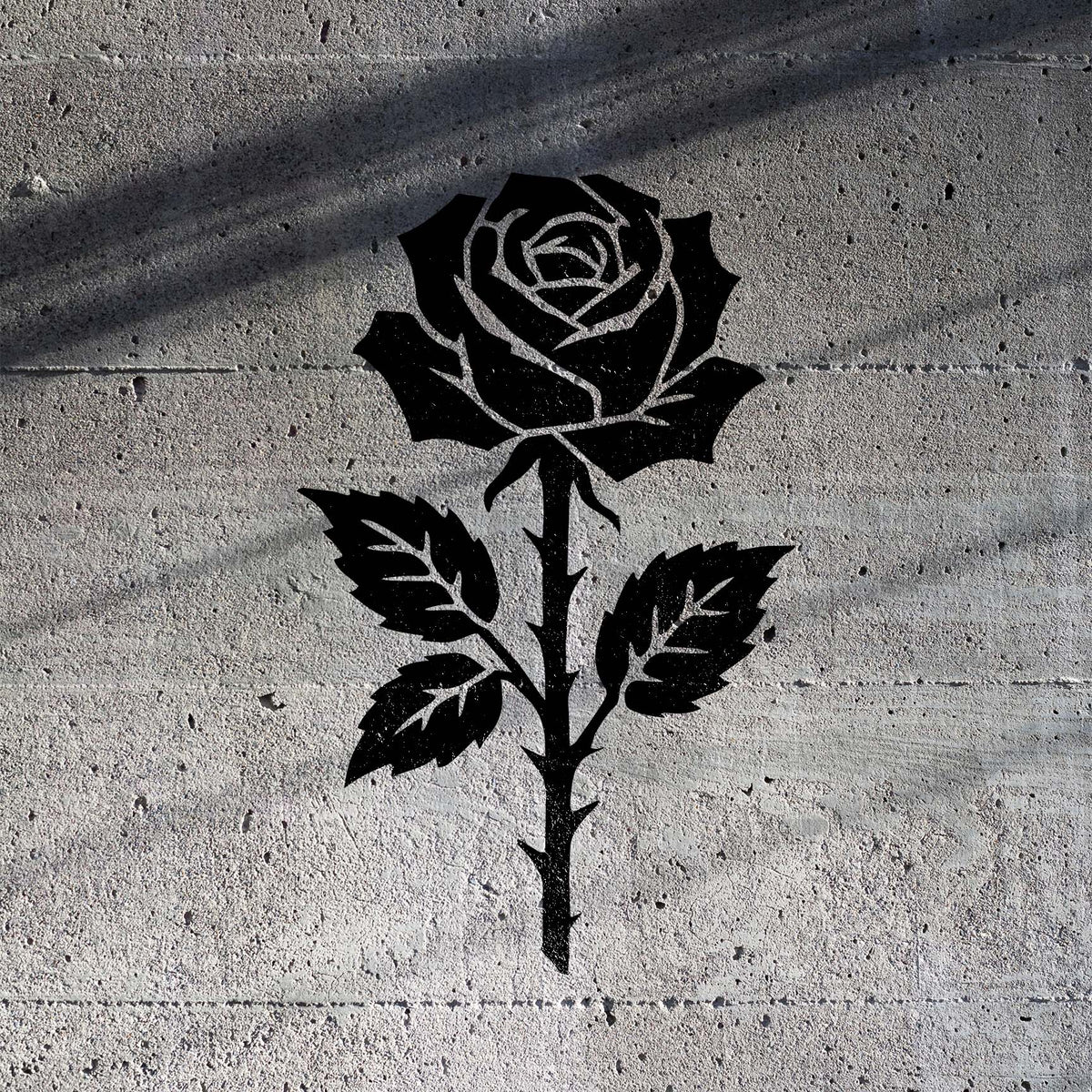 Thorned Rose Blossom Stencil in black painted on a textured concrete wall with soft shadow lines.