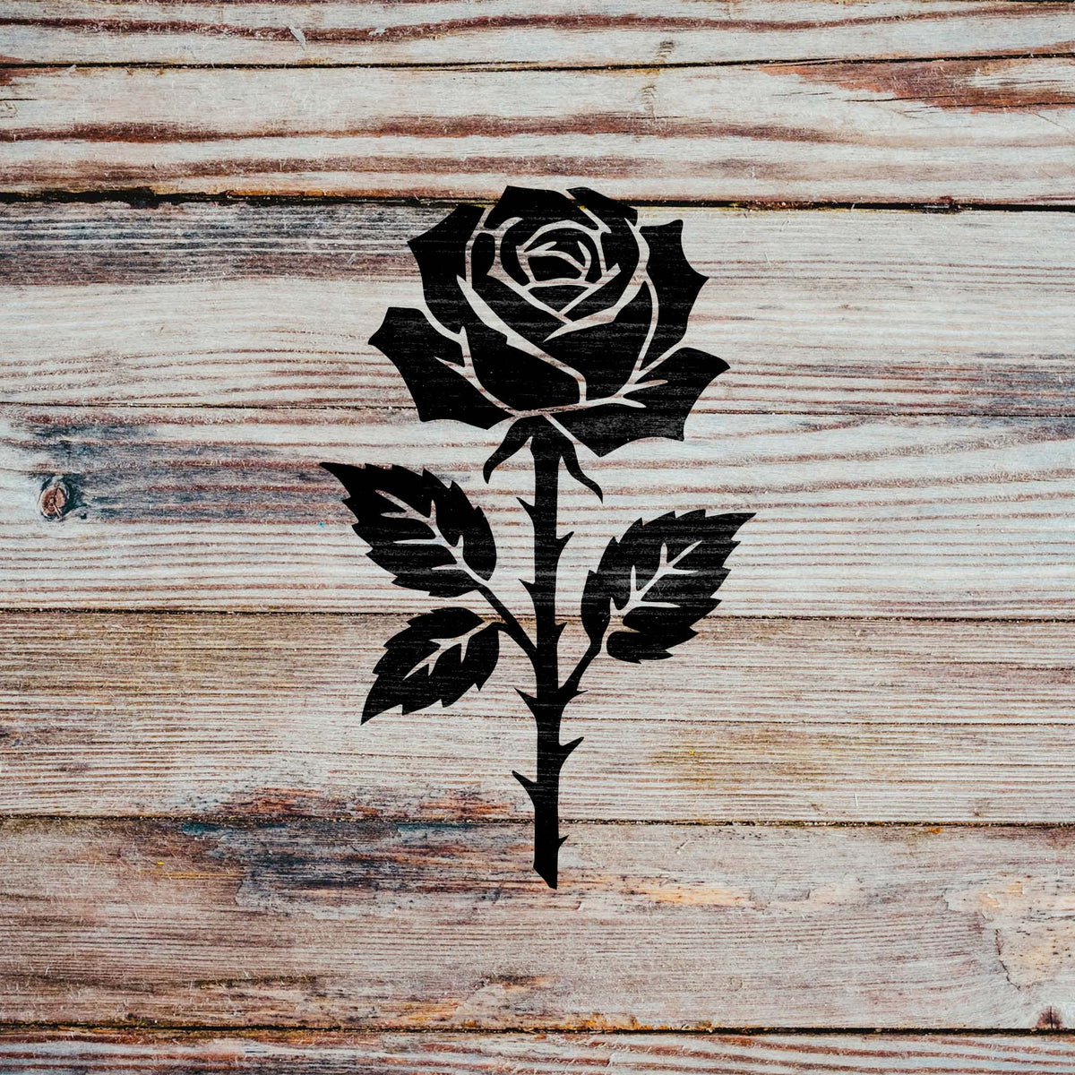 A bold black Thorned Rose Blossom Stencil stands out against a rustic light wood plank surface.