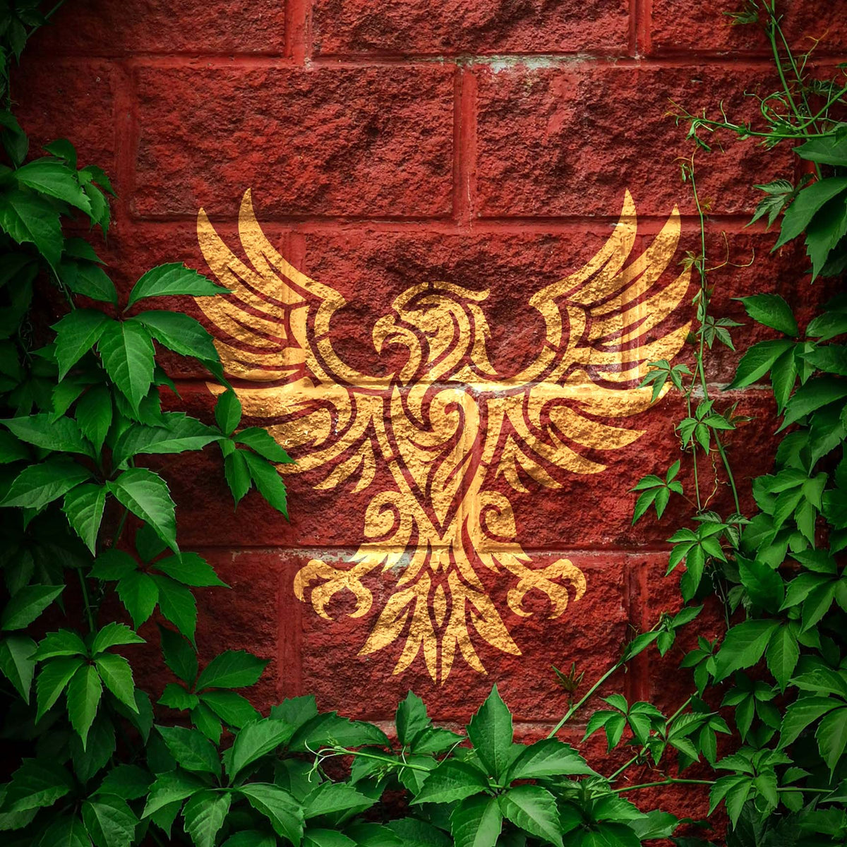 Gold Tribal Eagle Emblem Stencil artwork spreads across a rustic red brick wall framed by green vines.