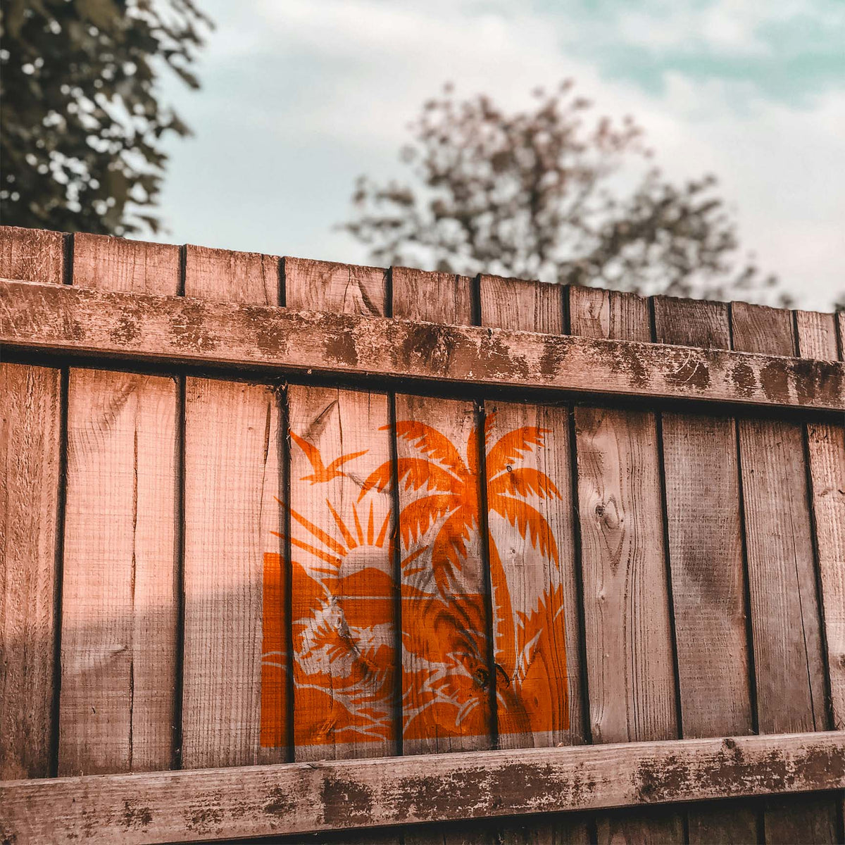 Orange Tropical Beach Sunset Stencil paints a palm, rolling surf and sun across a rustic wood fence