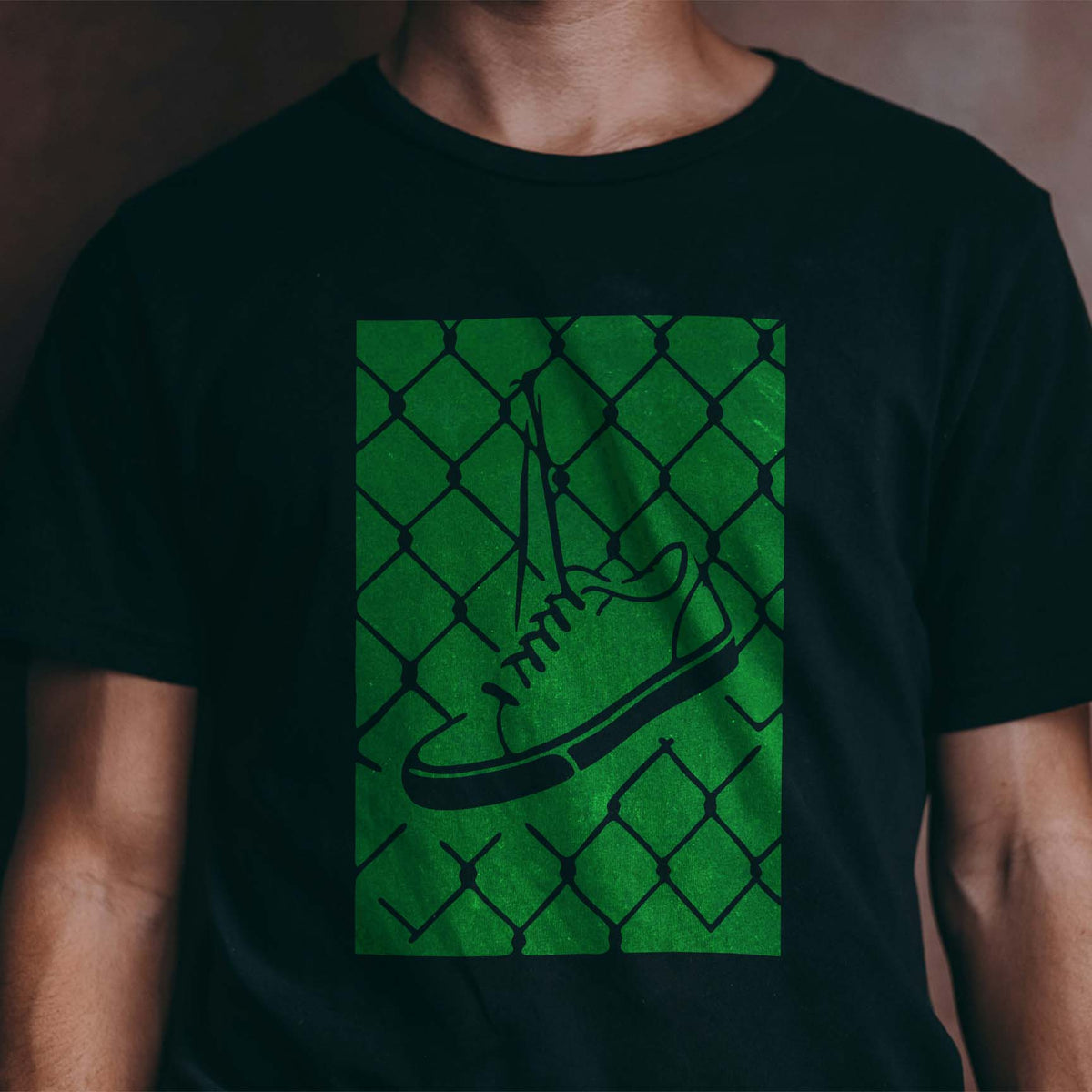 A green Urban Hanging Sneaker Chain Link Fence Stencil design stands out on a black T-shirt.
