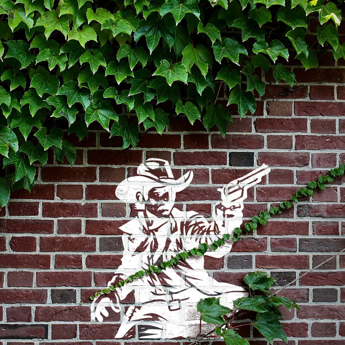 Amid climbing ivy, the Western Cowboy Revolver Stencil shows a white cowboy aiming a gun on a brick wall.